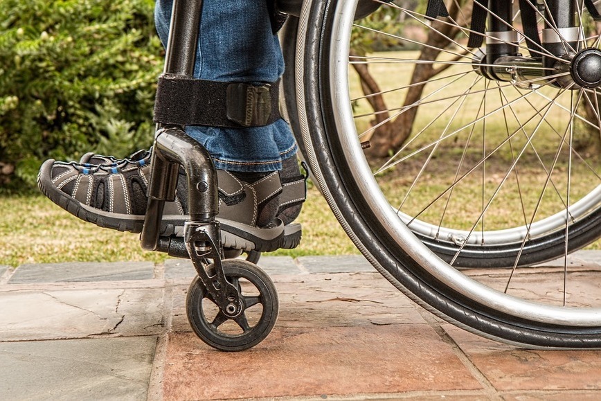 Zdjęcie przedstawia zbliżenie na wózek inwalidzki na którym ktoś siedzi, widać koło wózka i stopy osoby na wózku.