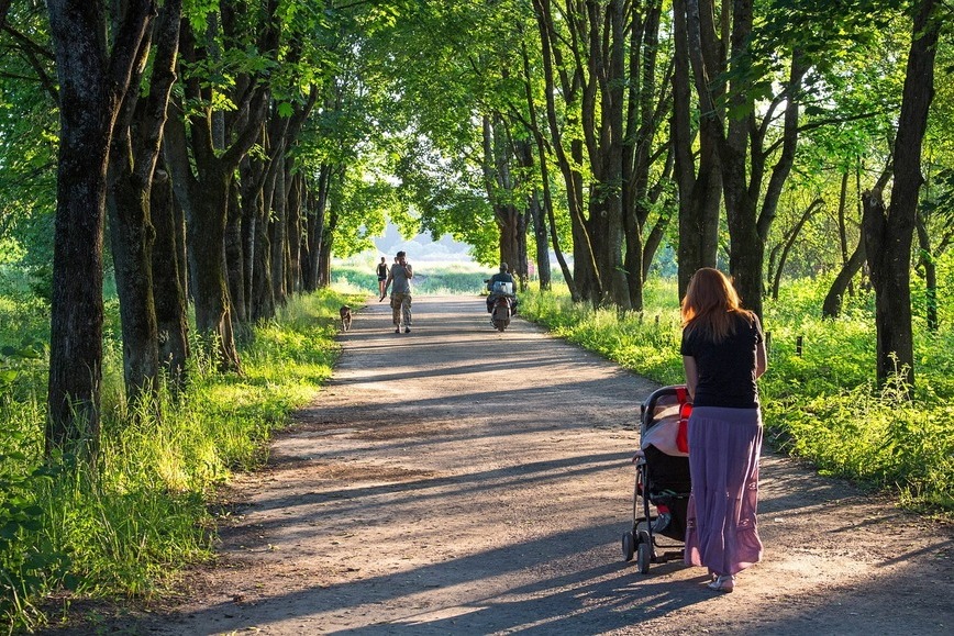 Zdjęcie przedstawia kobietę na spacerze z wózkiem w parku. W oddali widzać inne osoby.