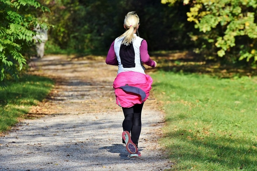 Zdjęcie przedstawia kobietę biegnącą w parku polną drogą.