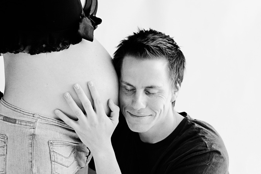 Czarno białe zdjęcie na którym mężczyzna przykłada ucho do brzucha kobiety w ciąży, jest zamyślony, ma zamknięte oczy.